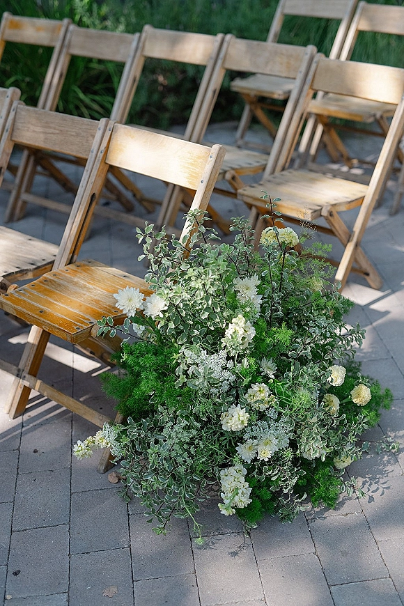 Ceremony seating with outdoor ceremony chairs lined on a stone paver patio, featuring white flower and greenery aisle florals in a garden setting