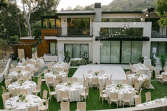Outdoor reception setup with round tables in white linens, upholstered chairs, and floral centerpieces beneath string lights on a lawn patio by a modern house