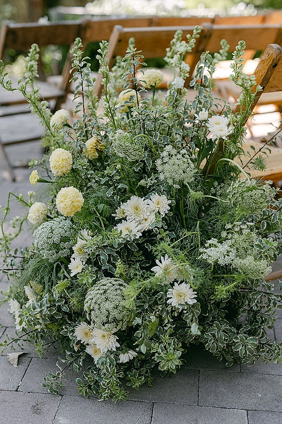Ceremony aisle flowers in grounded aisle florals with greenery and white blooms, with pale yellow accents beside wooden chairs on a stone patio