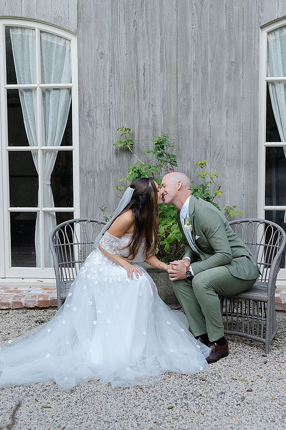 Wedding kiss portrait of bride and groom kissing while seated on wicker chairs, holding hands, in front of wood siding and white windows