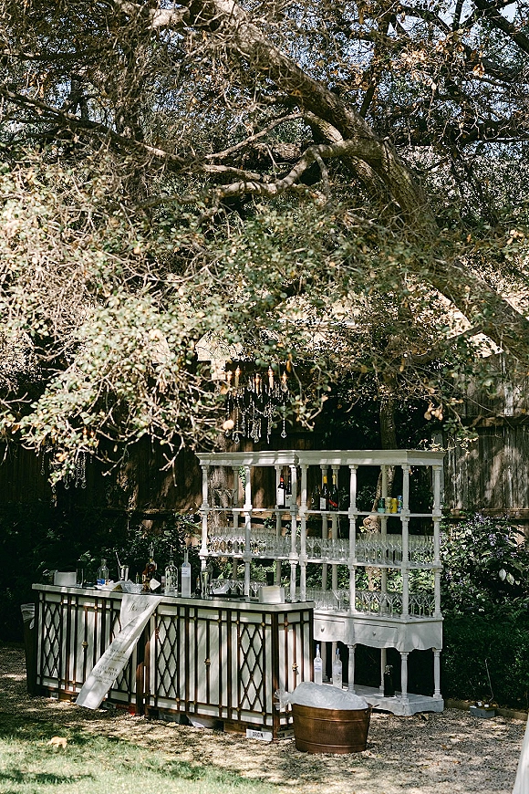 Outdoor wedding bar with white shelving, liquor bottles and glassware, topped by a crystal chandelier under a leafy garden canopy