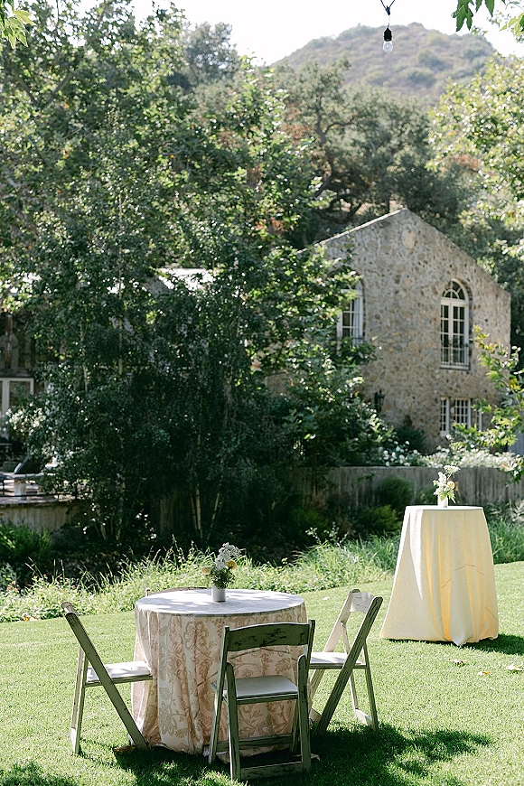 Outdoor reception setup with round tables and cocktail tables, small floral centerpiece, and string lights on a grassy lawn by a stone building