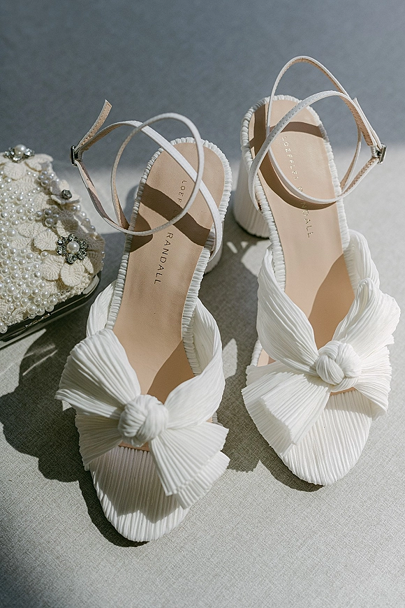 Bridal shoes and white bow heels with ankle straps beside a pearl beaded clutch on linen fabric, lit by soft sunlight and shadows
