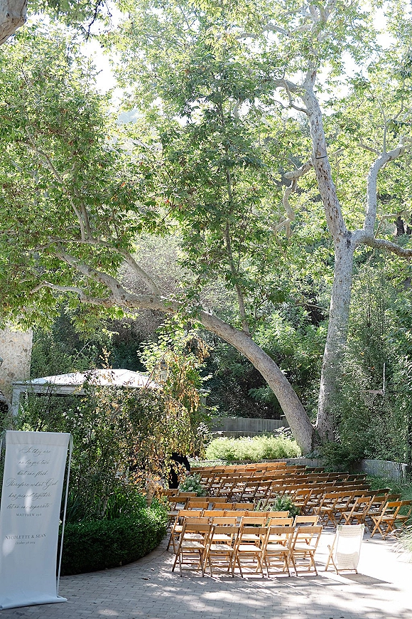 Outdoor ceremony setup with wood folding chairs lining an aisle runner and ceremony signage on a stone patio under trees on a hillside