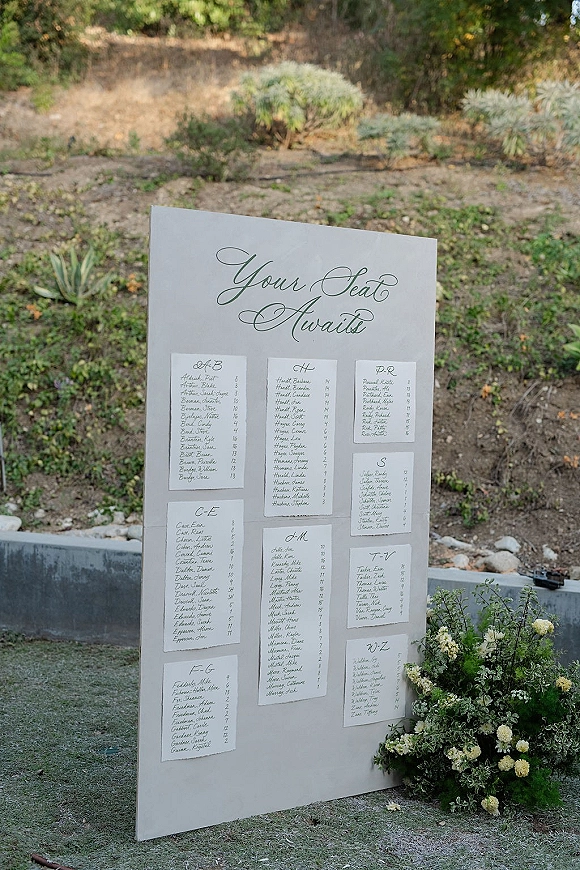 Wedding seating chart with calligraphy seating chart cards on a display board, framed by white flowers and greenery on a garden path