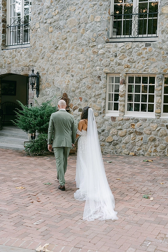 Couple portrait of bride and groom walking away, holding hands as her long wedding veil train trails across brick pavers by stone windows
