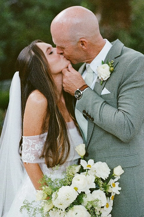 Wedding kiss portrait of bride and groom kissing, groom cradling her face, veil and white bouquet against lush outdoor greenery
