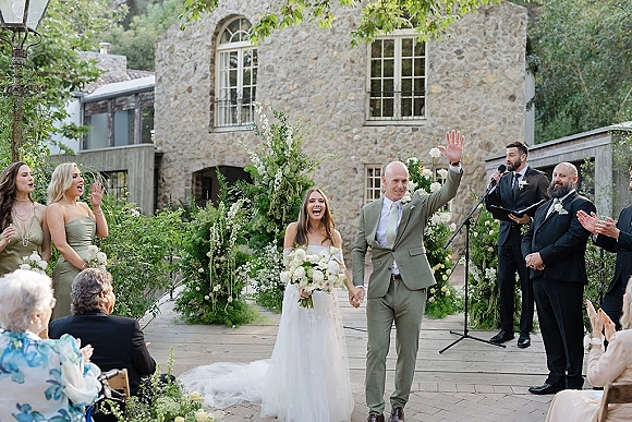 Recessional moment as bride and groom walk aisle holding hands, bouquet raised; guests cheer on a wooden deck by a stone building