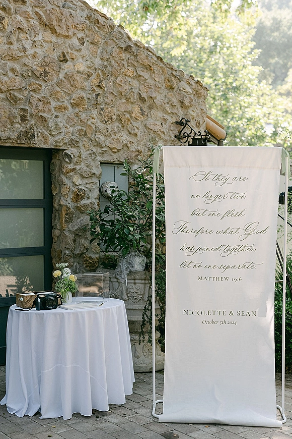 Wedding welcome sign with calligraphy wedding welcome sign on a stand, beside a white-clothed table with flowers, card box and instant camera.