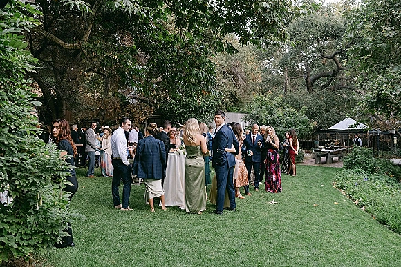 Wedding cocktail hour with guests mingling around linen cocktail tables, holding drinks on a garden lawn beneath large trees and umbrellas