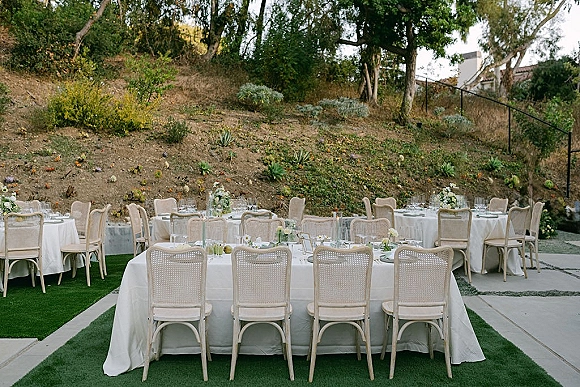 Reception tablescape for an outdoor wedding reception with white linens, cane back chairs, floral centerpieces and candles on a hillside patio