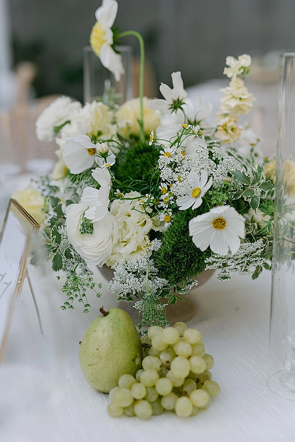 Wedding centerpiece with a white and green centerpiece in tall glass cylinder vases, with pears, grapes, and a table number on white linen