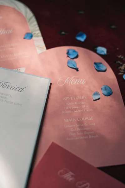 Wedding menu card with arched pink design and white calligraphy, styled with blue flower petals on a dark wood flat lay background