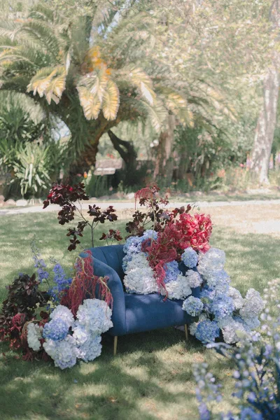 Wedding lounge decor with an outdoor wedding lounge blue velvet sofa surrounded by hydrangeas, red flowers, and greenery on a sunny garden lawn with palm trees