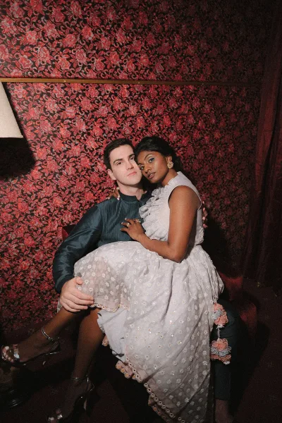 Couple portrait of bride on groom lap in a dim lounge, her ruffled wedding dress and gold earrings against red floral wallpaper
