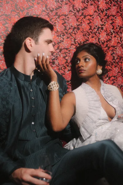 Couple portrait of bride touching groom’s face, her statement earrings and embroidered white lehenga against a red patterned tapestry backdrop