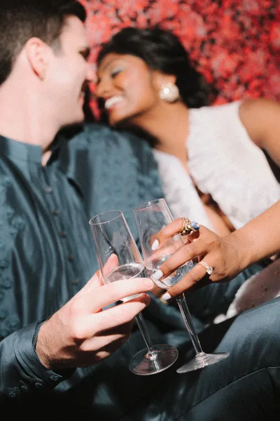 Wedding toast moment as newlyweds clink champagne flutes, bride’s engagement ring and white nails visible against a red floral wall backdrop