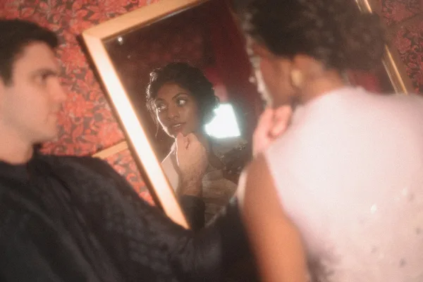 Bridal makeup touch-up as the bride getting ready looks in a mirror while a makeup brush applies soft glam in window light by patterned wallpaper