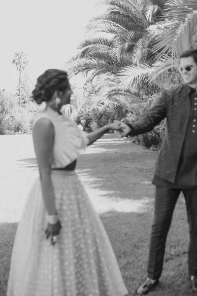Couple portrait of bride and groom holding hands, walking on a garden lawn with palm trees; bride’s statement earrings and groom sunglasses stand out