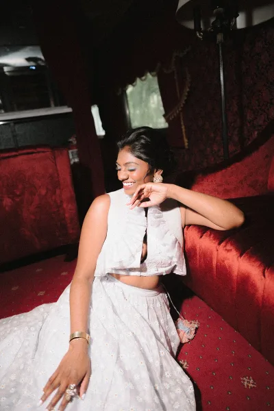 Bridal portrait of a bride sitting on floor in a white embroidered two-piece outfit, statement earrings and updo, by red velvet seating in dim window light