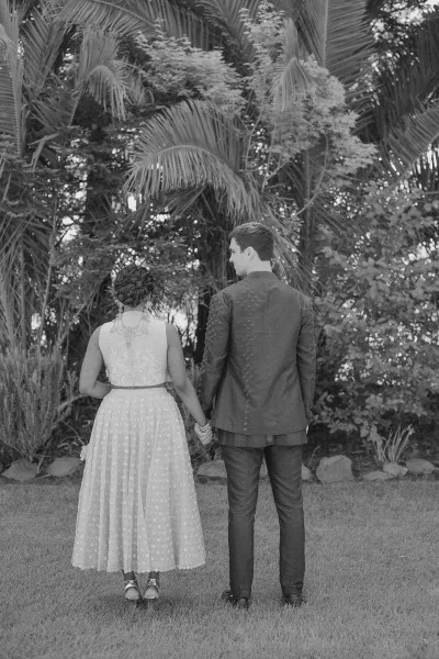 Couple portrait of bride and groom from behind holding hands, her lace dress accented by jewelry, walking past palm trees and rocks