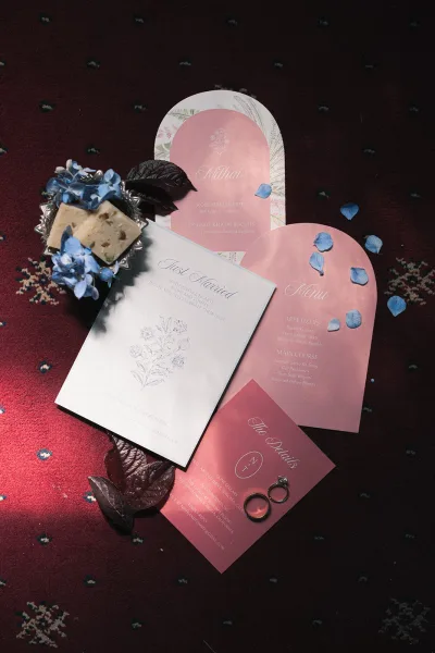 Wedding invitation suite wedding stationery flat lay with pink arched cards, menu and detail card, wedding rings and blue petals on patterned carpet