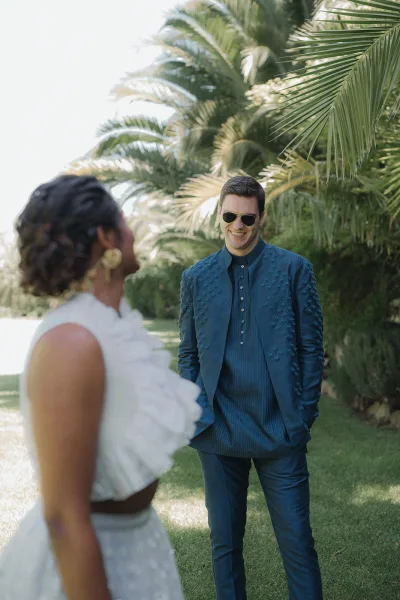 First look moment as bride in a ruffle neckline gown and statement earrings meets groom in sunglasses and embroidered sherwani on a palm-lined lawn