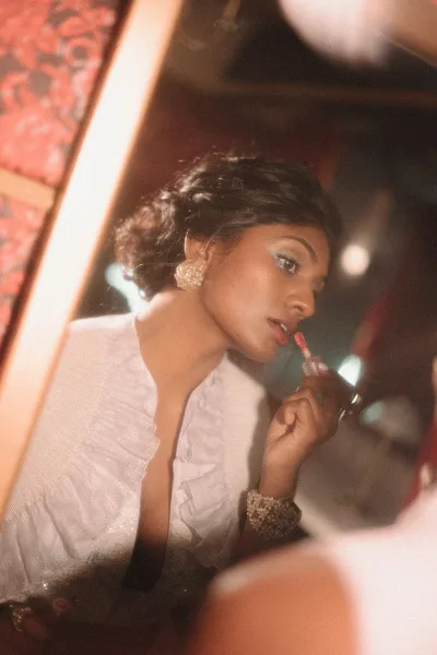 Bridal makeup as the bride applying lip gloss in a mirror, wearing statement earrings and an embroidered robe in a dim, warm-lit room