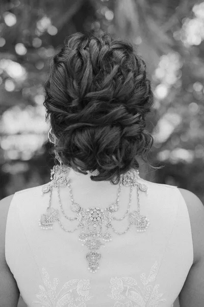 Bridal updo with low chignon wedding hair, showing lace-embroidered strapless gown and statement necklace against soft tree bokeh backdrop