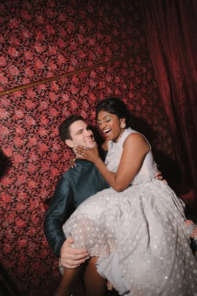 Couple portrait of bride on groom lap, laughing as she holds his face, showing engagement ring and earrings against red floral wallpaper backdrop