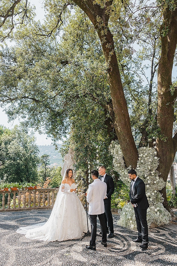 Wedding vows as bride reads from paper cards beside groom in tuxedo under a white floral arch on a garden terrace with trees