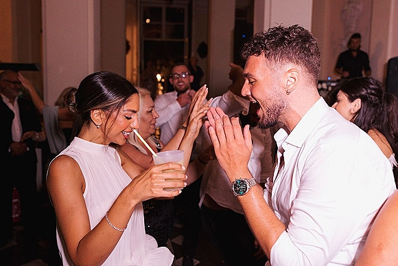 Wedding dance floor reception dance party with guests clapping as couple dances; cocktail cup with straw in foreground near the DJ booth