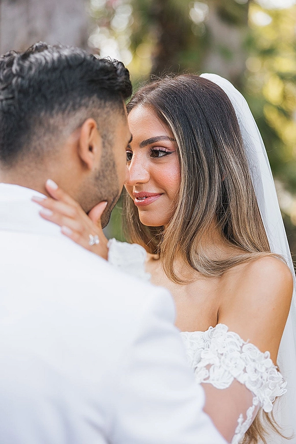 Wedding couple portrait of bride and groom close up, forehead to forehead as she holds his face, veil and lace dress in soft greenery bokeh