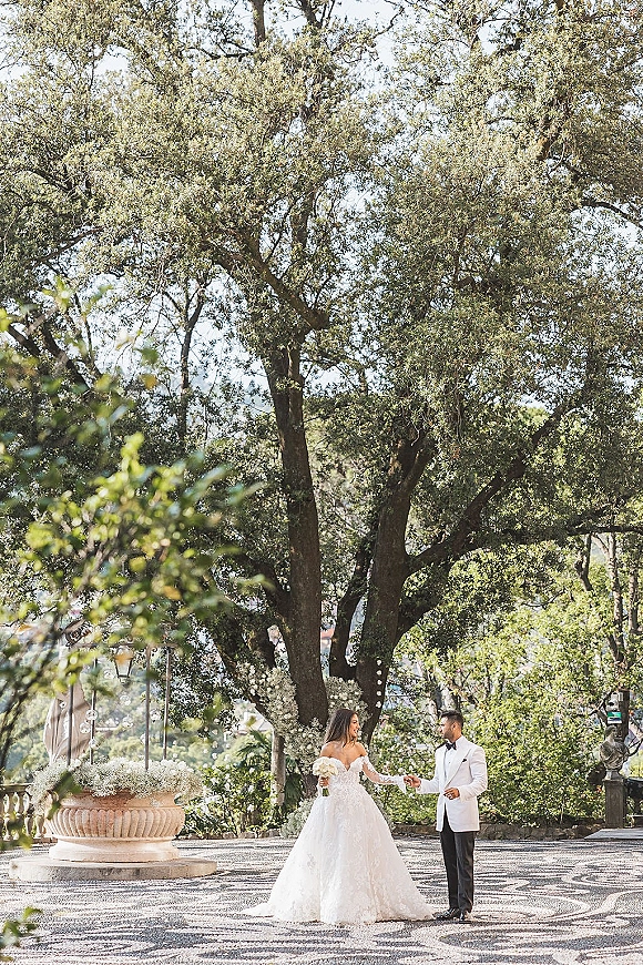 Couple portrait outdoors, bride and groom holding hands on a stone terrace with garden trees, urn planter and string lights behind