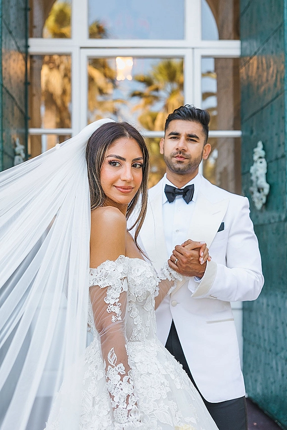 Couple portrait of bride and groom posing, bride looking over shoulder in long veil and lace sleeves beside glass doors with green shutters