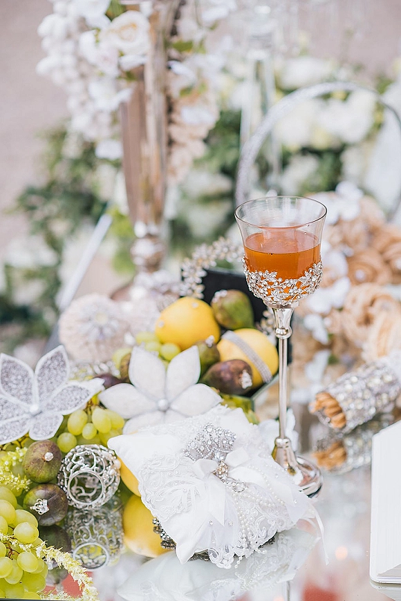 Wedding tablescape with wedding table styling of wine glass, amber drink, lemons, grapes and figs among white florals and jeweled décor outdoors