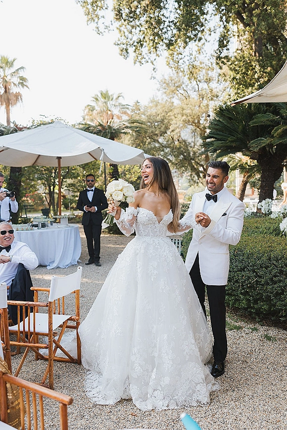 Couple portrait of bride and groom laughing as she twirls her lace dress with a white rose bouquet on a garden patio with umbrellas