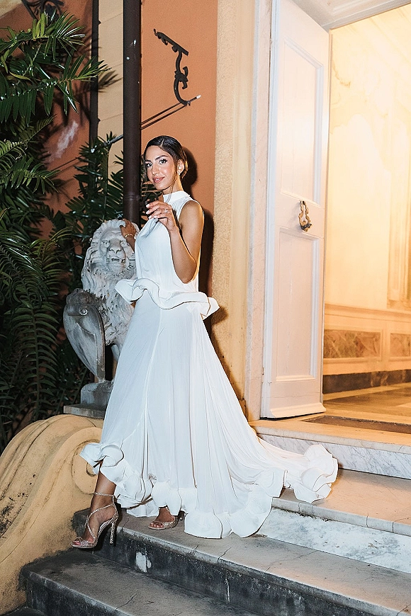 Bridal portrait of a bride on stairs in a white high-neck gown with ruffle hem and train, holding her dress on stone steps by a doorway