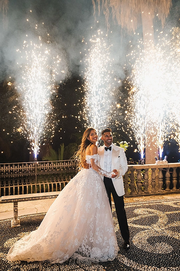 Couple portrait of bride and groom holding hands during a sparkler exit wedding, framed by smoke and fireworks on a stone terrace at night