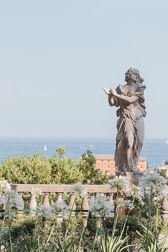 Garden statue on a stone balustrade terrace with white flowers and greenery, overlooking a blue ocean with sailboats and coastline buildings