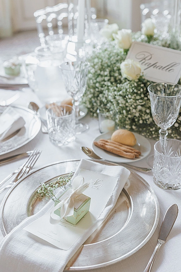 Reception tablescape with wedding place setting on silver charger plates, crystal goblets, white linens, roses and baby’s breath in soft window light