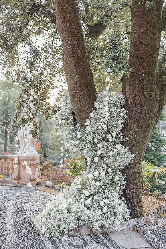 Wedding floral installation of white roses and baby's breath garlands wrapping a large tree, cascading with greenery over a stone patio