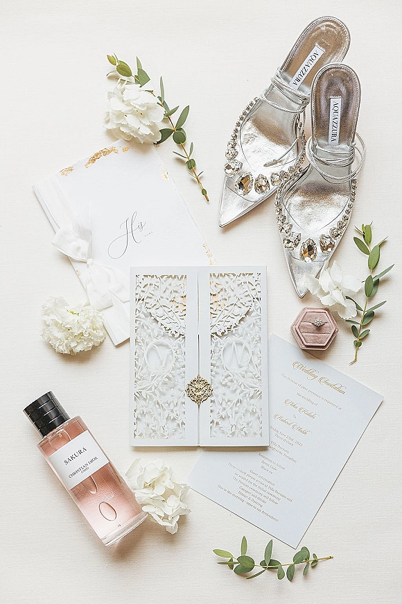 Invitation suite with laser cut wedding invitations, ribbon and wax seal, styled with ring box, jeweled shoes, perfume on white fabric backdrop