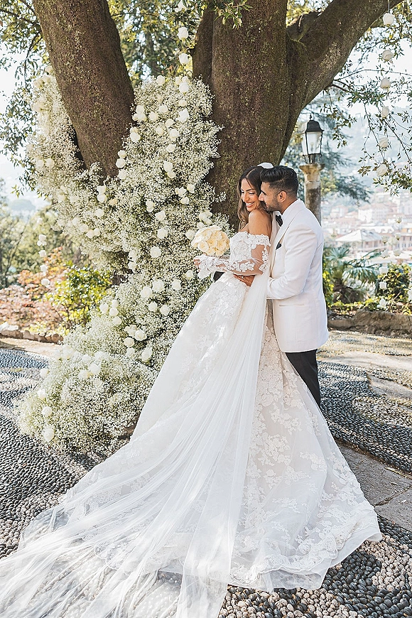 Couple portrait of bride and groom embrace beneath hanging flowers by a large garden tree, her lace gown and veil trailing as she holds a white rose bouquet