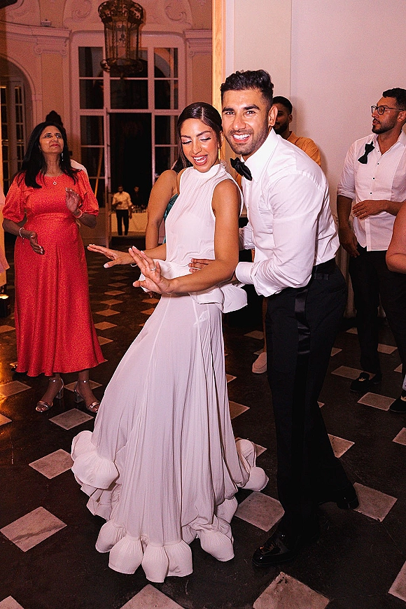 Wedding reception dancing as bride in a high-neck gown and groom in bow tie spin on a tiled ballroom floor under chandelier, guests watching