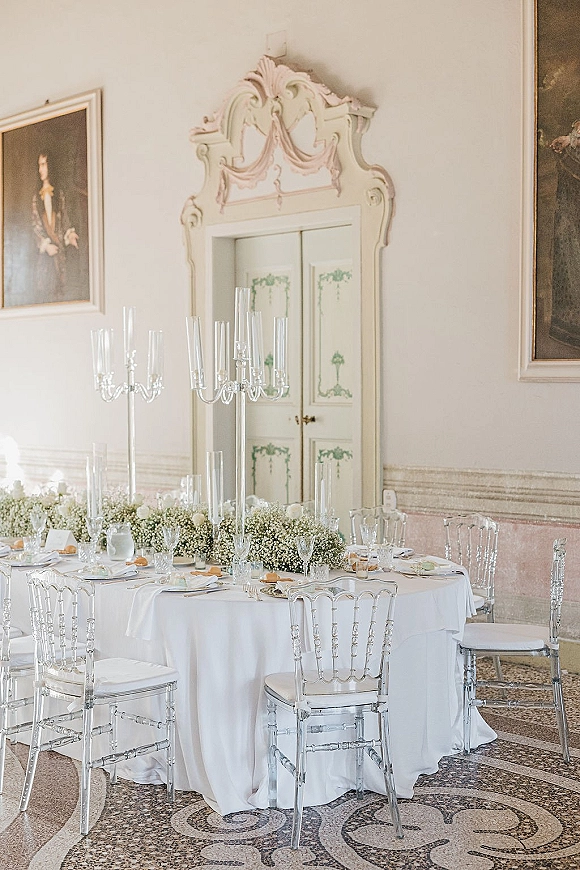 Reception tablescape with round wedding table setup, tall glass candelabras and white roses on baby's breath garland in ornate hall