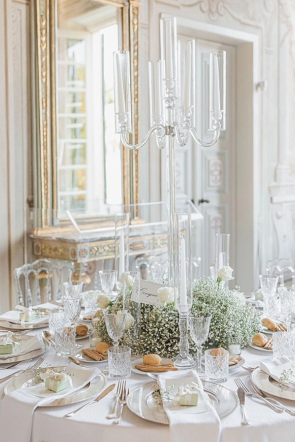 Reception tablescape with wedding table centerpiece featuring tall glass candelabra, white flowers and crystal goblets in an ornate room with gilded mirror