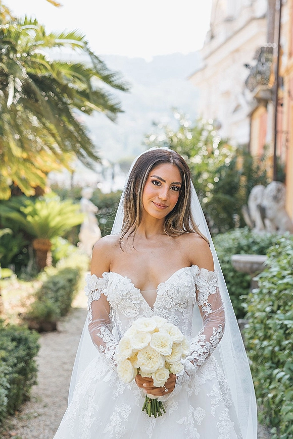 Bridal portrait of a bride in a lace wedding dress holding a white rose bouquet, cathedral veil trailing along a garden path with palms