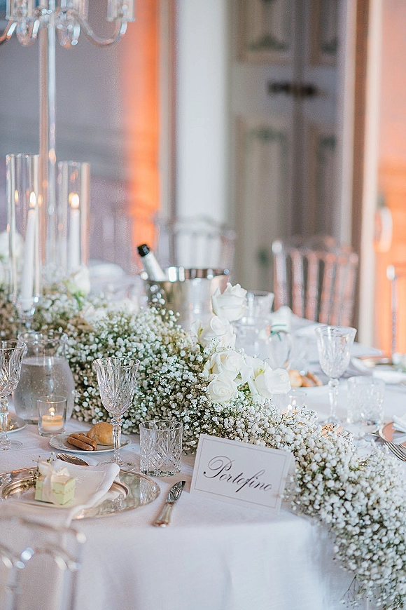 Reception tablescape with wedding head table decor, white rose centerpiece and baby’s breath garland, taper candles under a crystal chandelier in a banquet room