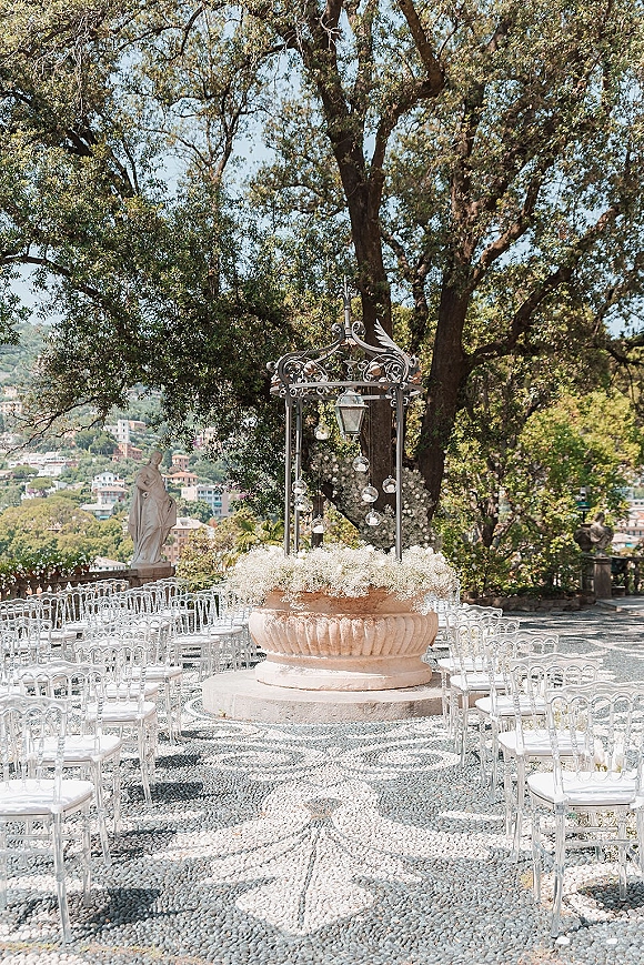 Ceremony setup for an outdoor wedding ceremony with clear chairs in a circular layout beneath a metal gazebo on a mosaic terrace overlooking hills